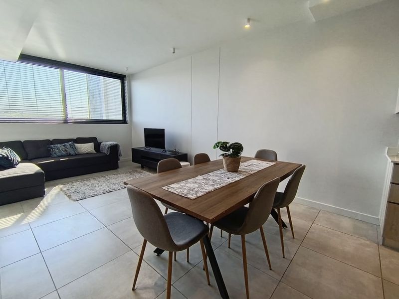 A bright open-plan living and dining area featuring neutral tiled flooring, recessed lighting, and large windows that provide ample natural light.
