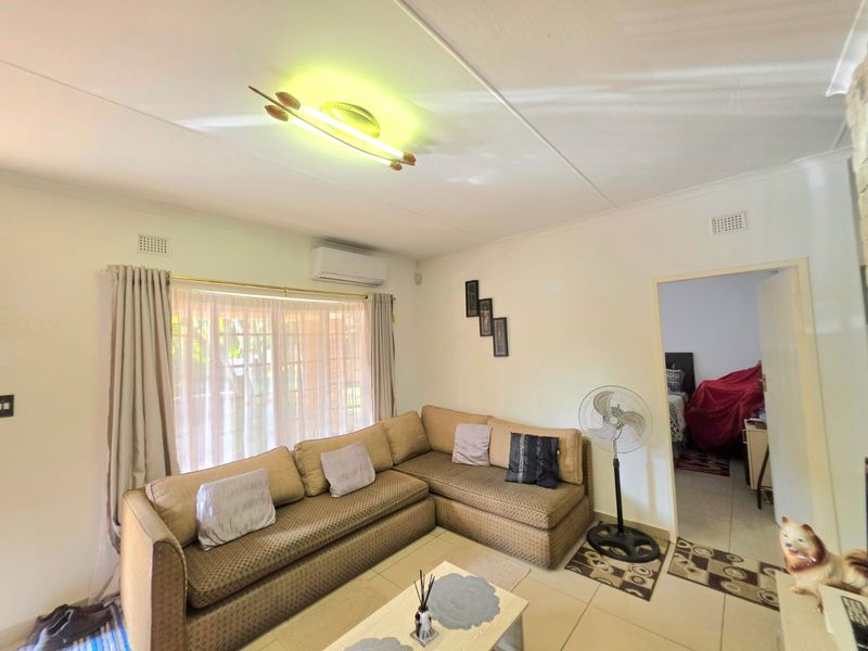 A living room featuring tiled flooring, a split-system air conditioner, and a large window with curtains, providing access to an adjacent bedroom.