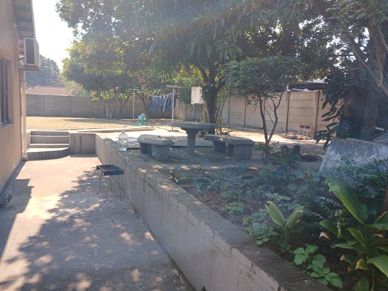 A spacious rear garden featuring a concrete patio area, retaining walls, mature trees, and a precast concrete boundary wall.