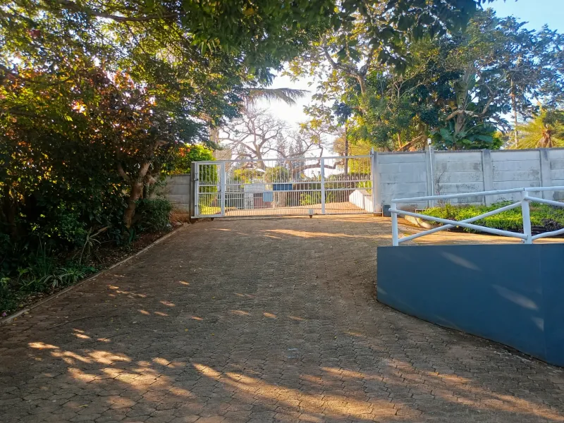 A paved driveway leading to a secure metal gate entrance, framed by mature trees and a perimeter wall.