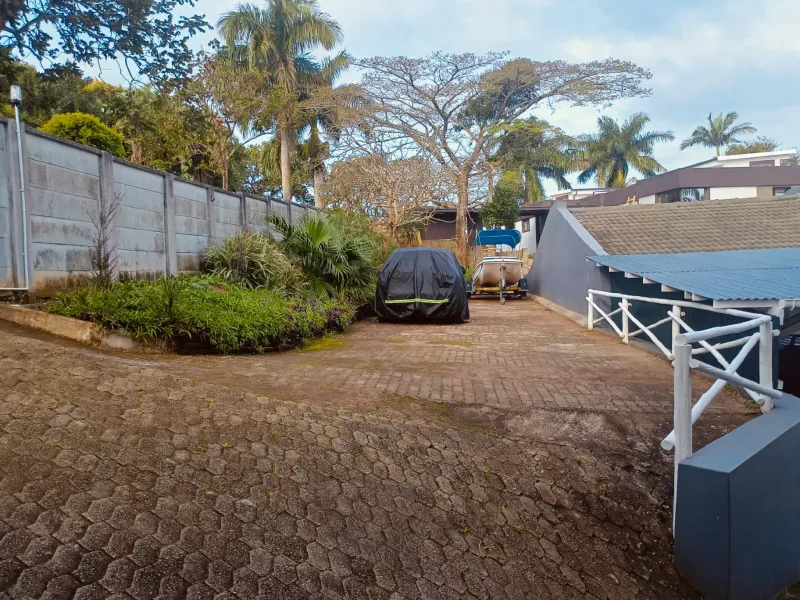 A paved driveway area featuring a precast concrete boundary wall, mature garden landscaping, and a covered structure with a corrugated metal roof.