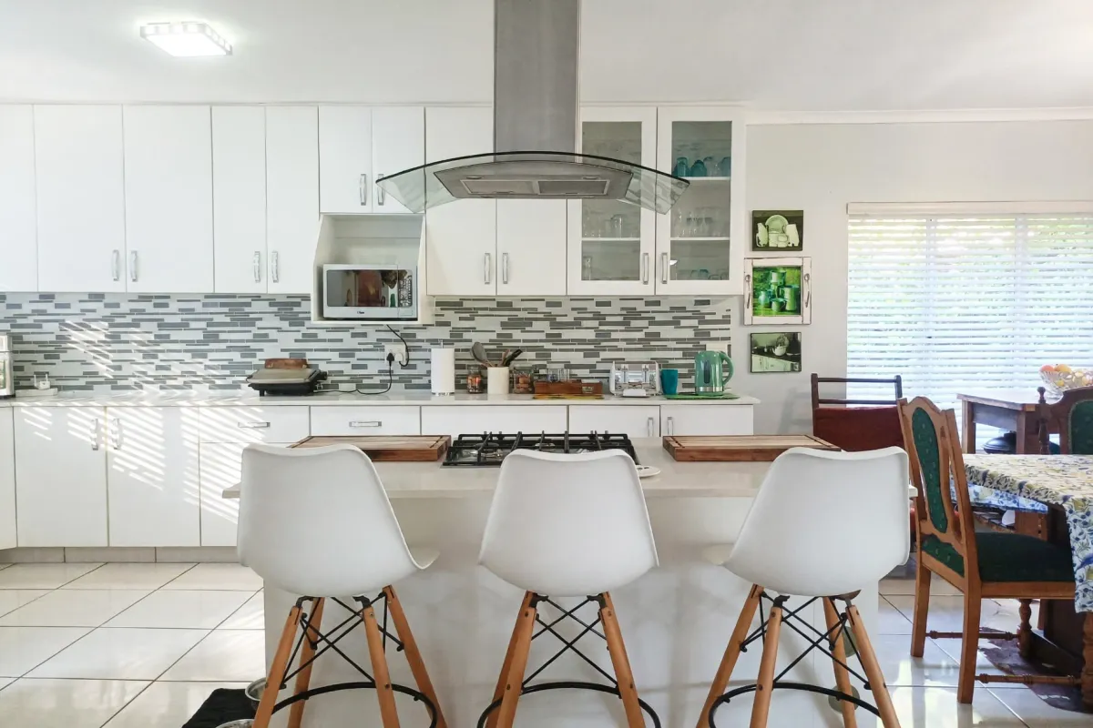 A bright, modern kitchen featuring white cabinetry, a central island with a gas hob, stainless steel extractor fan, and a stylish mosaic tile backsplash.