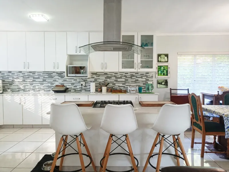 A bright, modern kitchen featuring white cabinetry, a central island with a gas hob, stainless steel extractor fan, and a stylish mosaic tile backsplash.