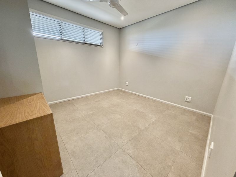A clean, empty bedroom featuring neutral tiled flooring, a ceiling fan, and venetian blinds, providing a versatile space ready for personalization.