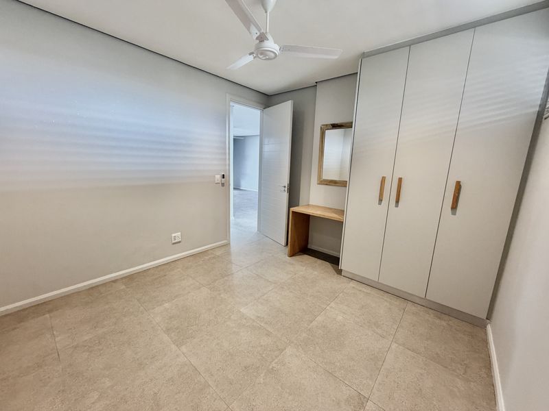 A clean, modern bedroom featuring neutral tiled flooring, a ceiling fan, and ample built-in cupboard space with an integrated vanity area.