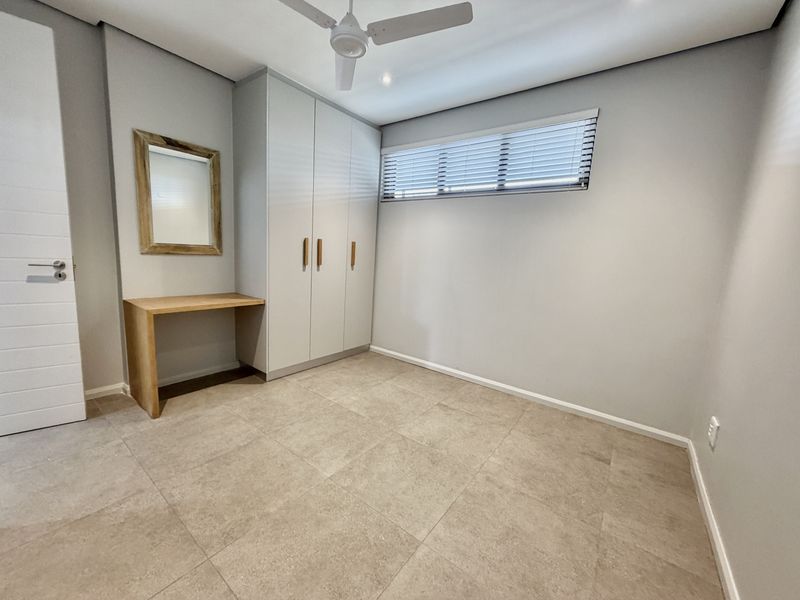 A clean, modern bedroom featuring neutral tiled flooring, built-in cupboards, a built-in desk, and a ceiling fan.