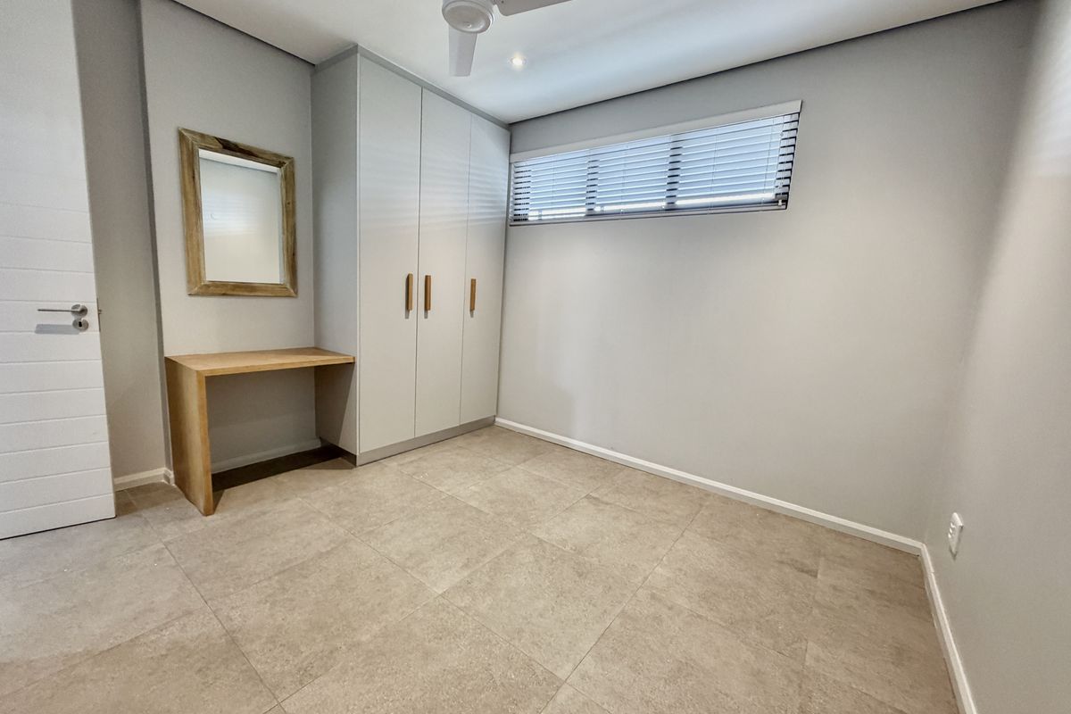 A clean, modern bedroom featuring neutral tiled flooring, built-in cupboards, a built-in desk, and a ceiling fan.