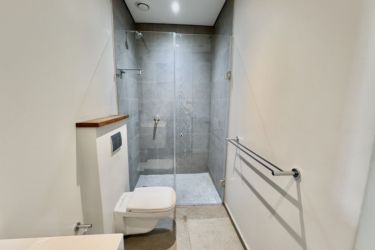 A modern, minimalist bathroom featuring a wall-hung toilet with a concealed cistern, a glass-enclosed shower, and neutral grey tiling.