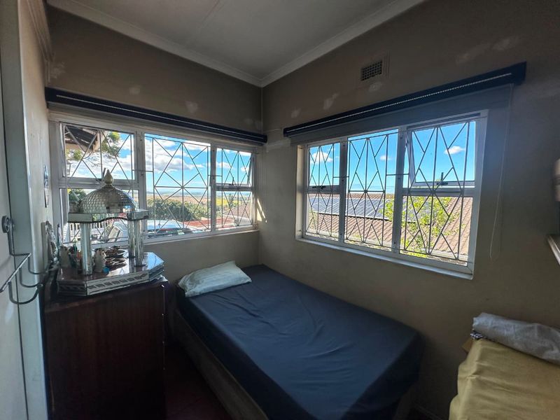 A compact bedroom featuring corner windows with security bars, roller blinds, and visible wall repair work requiring finishing.