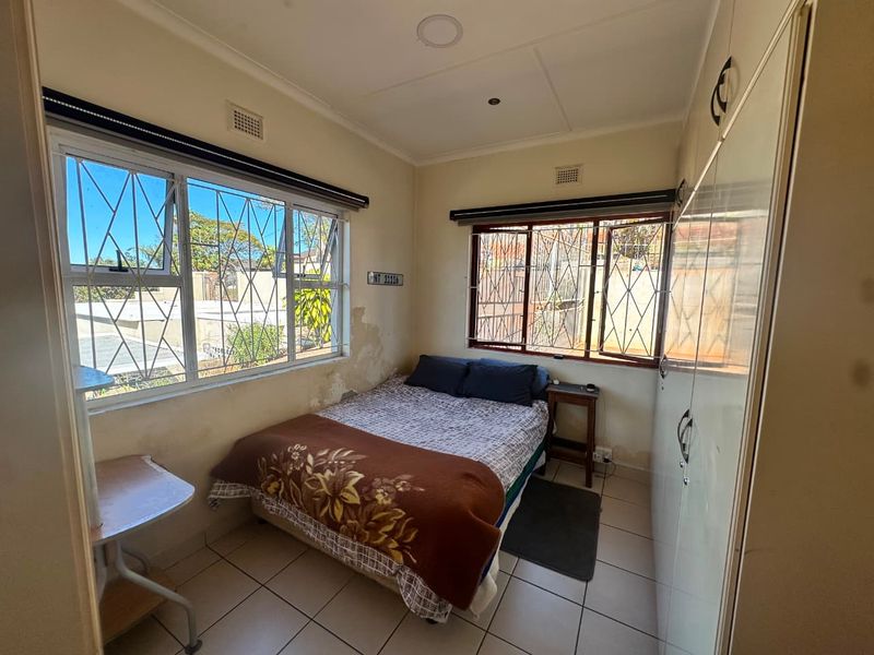 A compact bedroom featuring tiled flooring, built-in cupboards, and security bars on the windows, currently showing signs of wall surface deterioration.