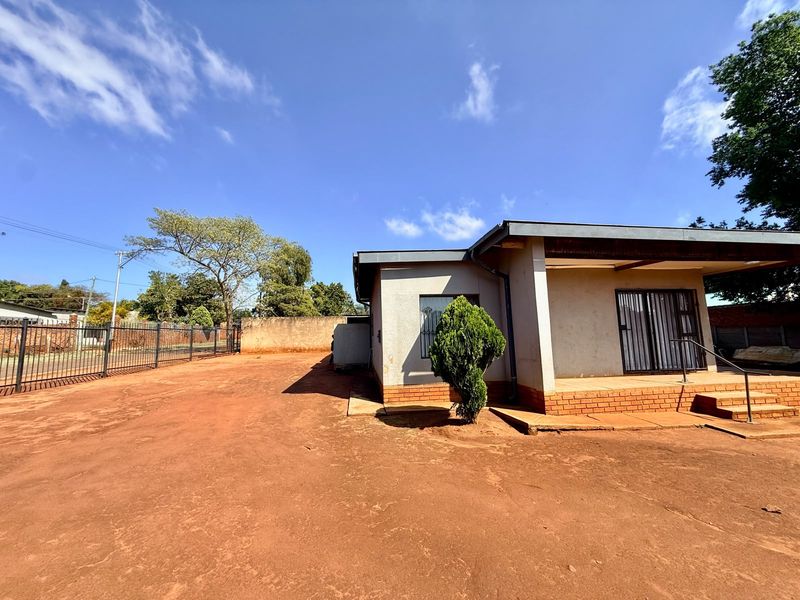 Front exterior view of the house featuring a large unpaved yard, metal perimeter fencing, and a covered entrance area with security gates.