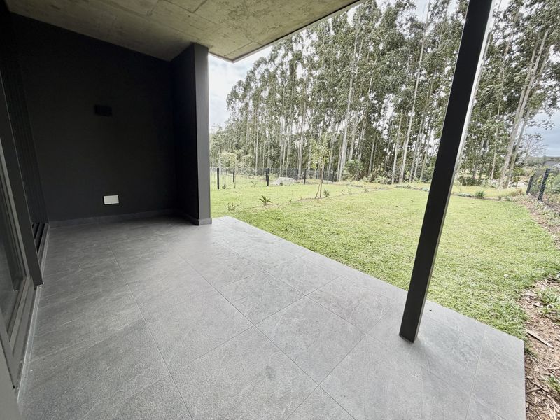 A modern covered patio featuring large format grey floor tiles, a concrete ceiling, and a support pillar, overlooking a private garden area.