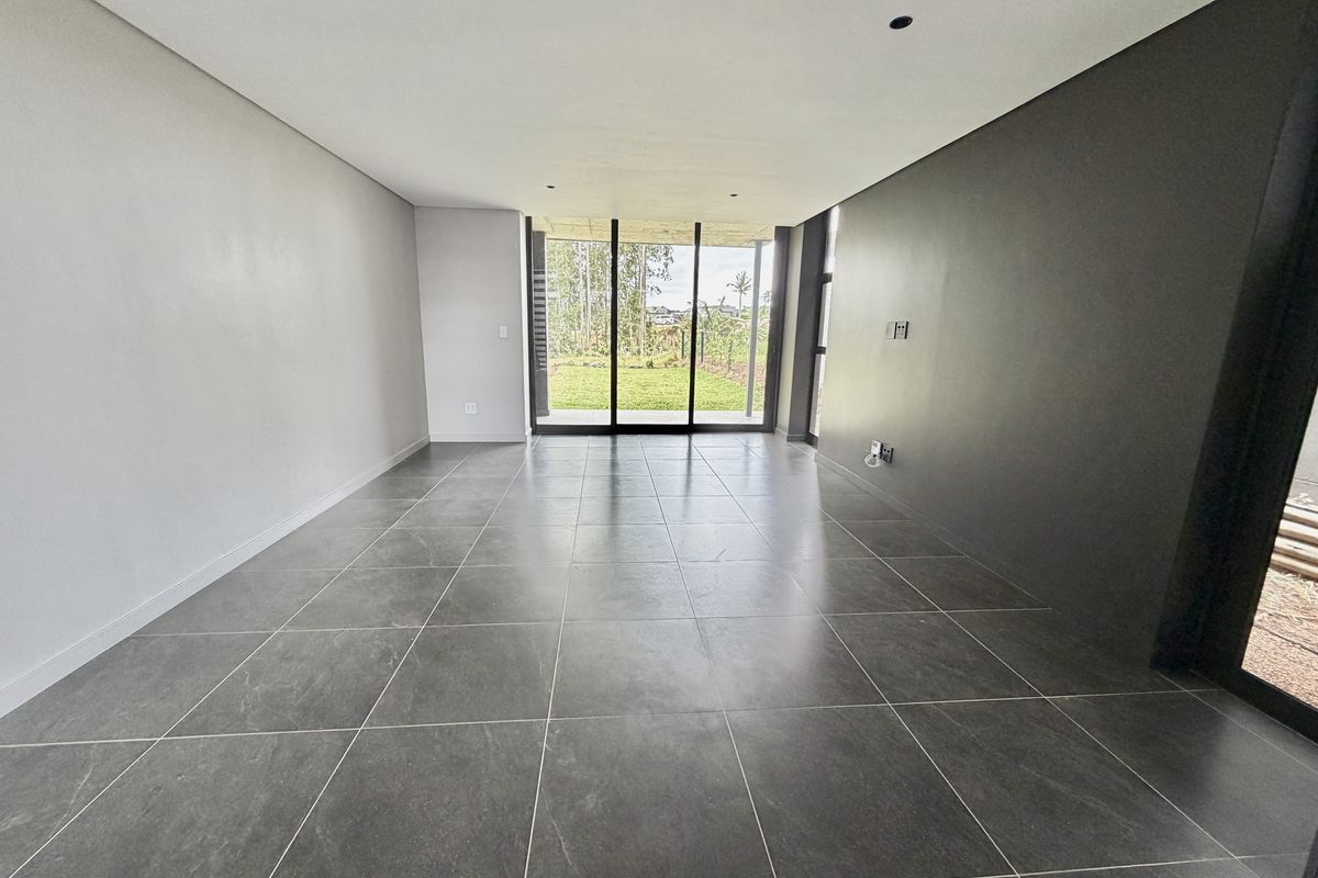 A modern, empty living area featuring large format dark floor tiles, recessed lighting, and sliding glass doors leading to a garden view.