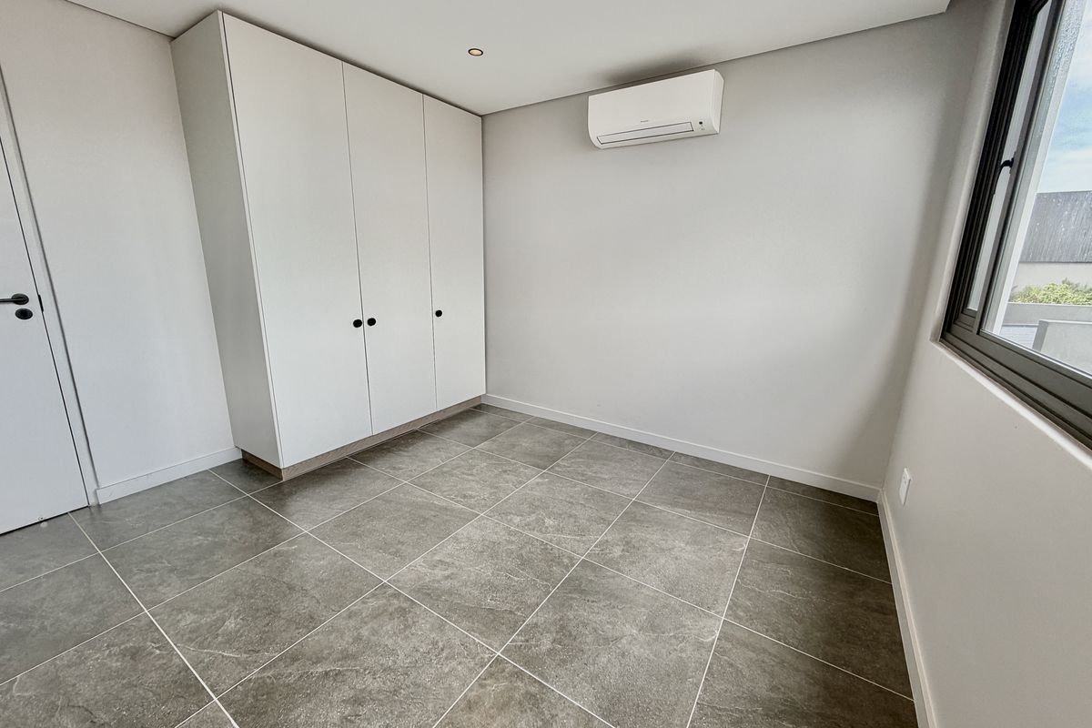 A clean, modern, and empty bedroom featuring neutral tiled flooring, built-in cupboards, and a wall-mounted air conditioning unit.