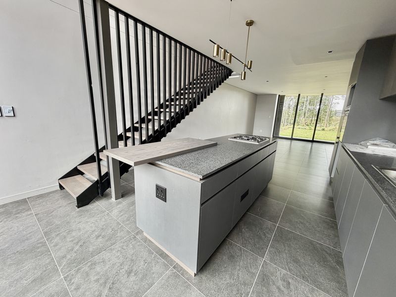 Modern open-plan kitchen featuring a central island with gas cooktop, sleek cabinetry, and a striking staircase, leading to a bright living area with garden views.
