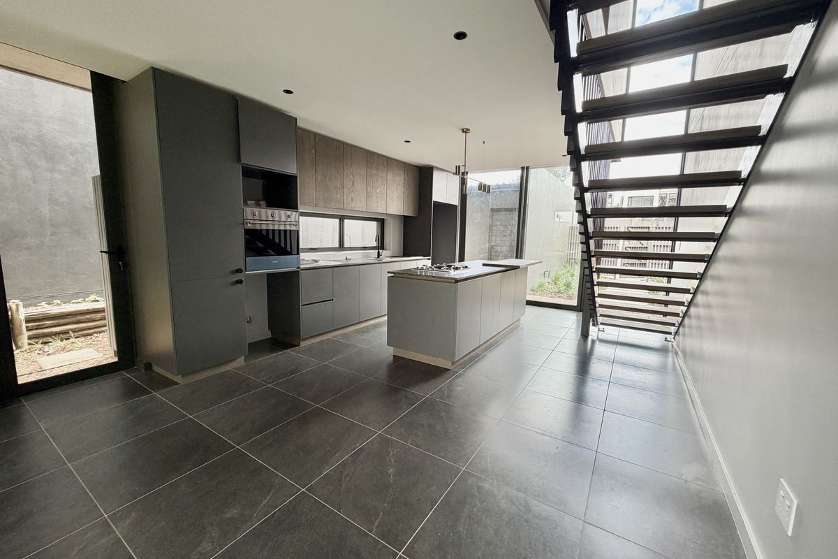 Modern kitchen featuring dark cabinetry, a central island with a gas cooktop, built-in oven, and large format floor tiles, adjacent to an open staircase.