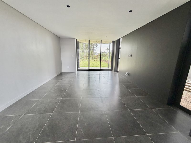 A modern, empty living area featuring large format dark floor tiles, recessed lighting, and sliding glass doors leading to a garden view.