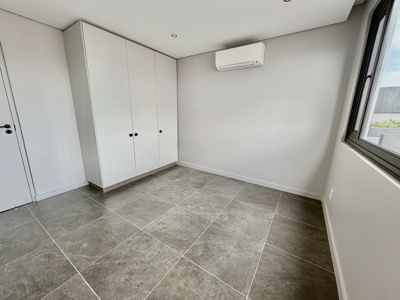 A clean, modern, and empty bedroom featuring neutral tiled flooring, built-in cupboards, and a wall-mounted air conditioning unit.