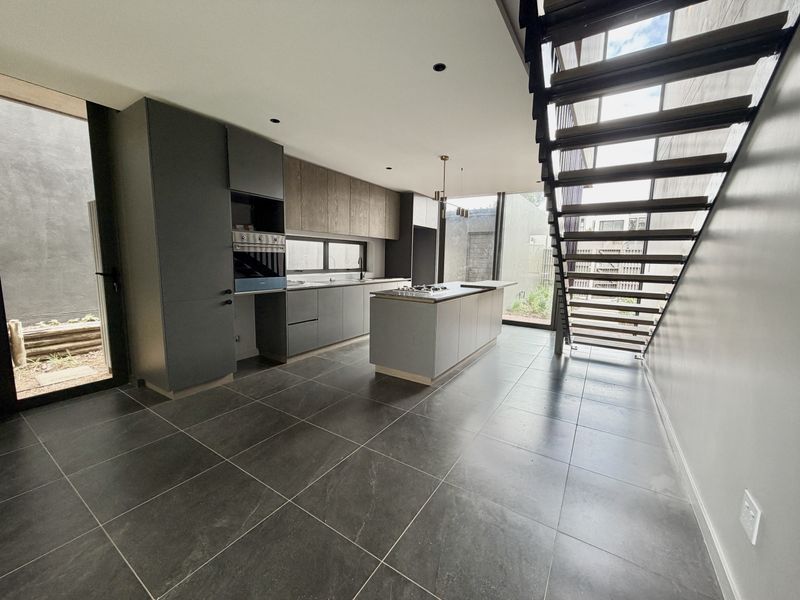 Modern kitchen featuring dark cabinetry, a central island with a gas cooktop, built-in oven, and large format floor tiles, adjacent to an open staircase.