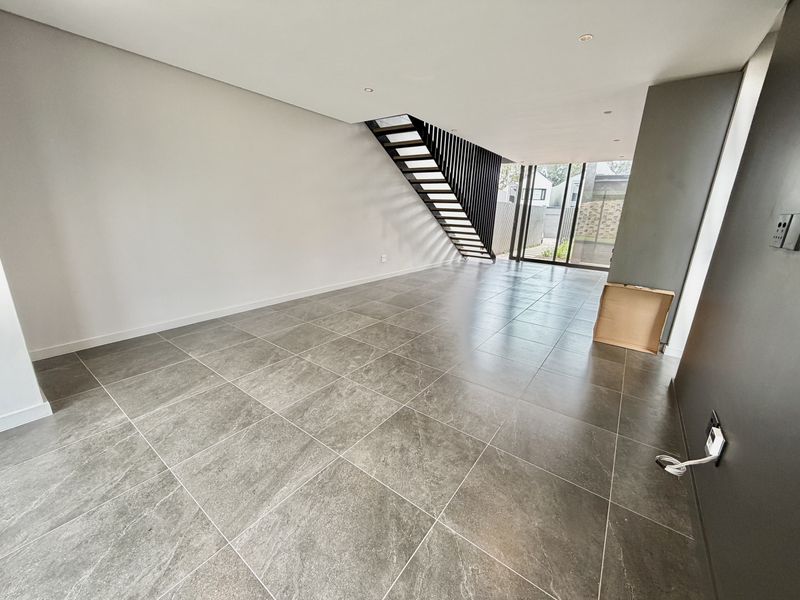 A spacious, modern open-plan living area featuring large-format grey floor tiles, a contemporary open-tread staircase, and sliding glass doors leading to the exterior.