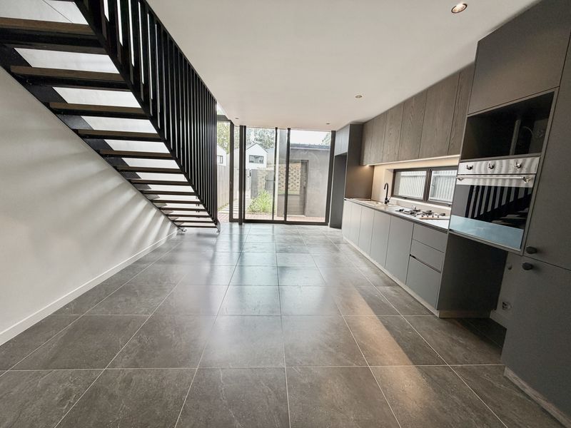 Modern open-plan kitchen and living area featuring sleek grey cabinetry, integrated appliances, large format floor tiles, and a contemporary staircase leading to the upper level.