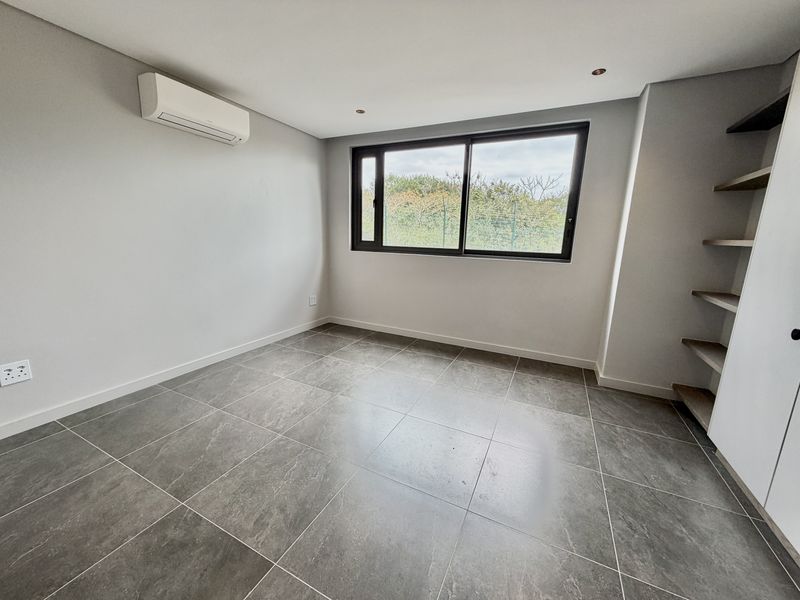 A bright, modern, and empty bedroom featuring large-format grey floor tiles, built-in shelving, and a wall-mounted air conditioning unit.