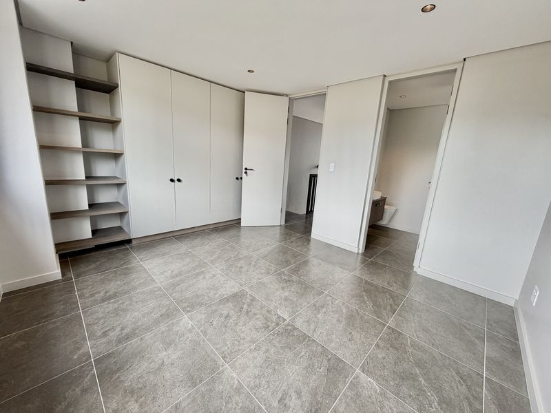 A modern, empty bedroom featuring large-format grey floor tiles, extensive built-in cabinetry with integrated shelving, and direct access to an en-suite bathroom.