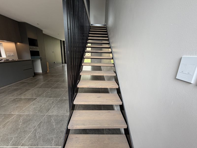 Modern open-plan living area featuring a contemporary timber staircase with metal balustrade, sleek tiled flooring, and a high-end kitchen with integrated appliances.