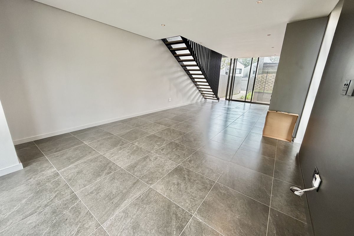 A spacious, modern open-plan living area featuring large-format grey floor tiles, a contemporary open-tread staircase, and sliding glass doors leading to the exterior.