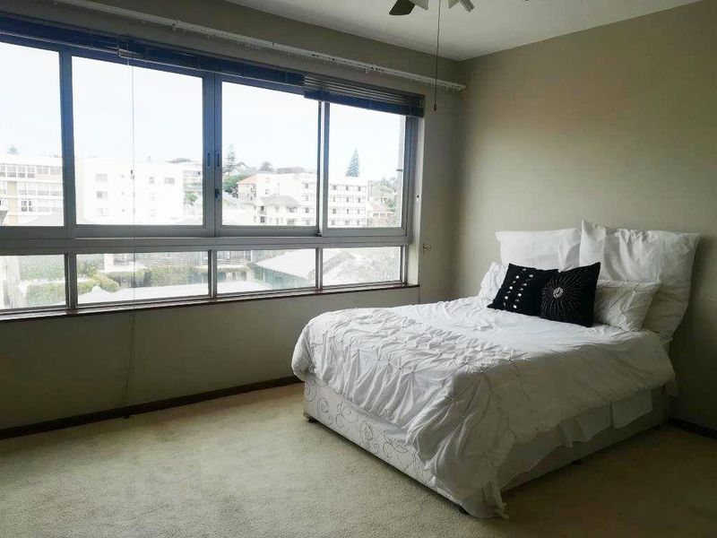A spacious bedroom featuring large windows that provide ample natural light and urban views, complemented by a ceiling fan and neutral carpeting.