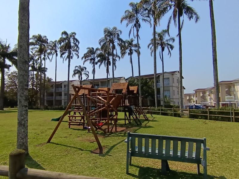 A well-maintained communal garden area featuring mature palm trees, manicured lawns, timber fencing, and a wooden playground structure within an apartment complex.