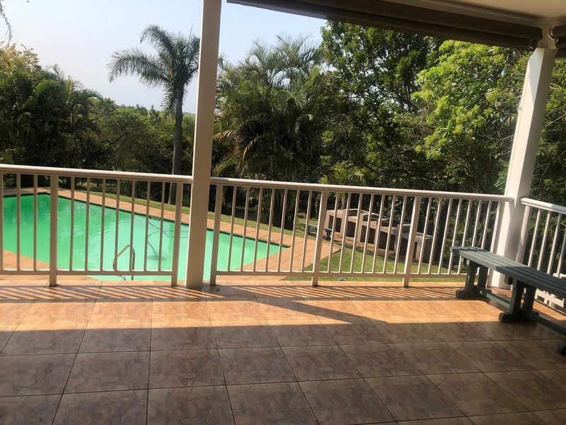 A covered patio featuring tiled flooring and a protective railing, overlooking a swimming pool and garden area with a built-in braai.