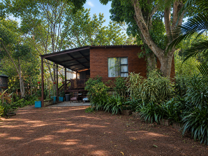 A charming log cabin cottage featuring a covered timber deck, surrounded by lush mature trees and established garden landscaping.