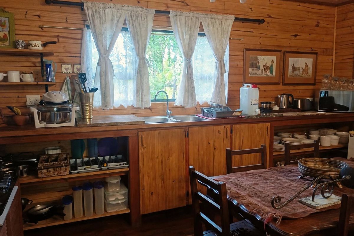 Rustic cottage kitchen featuring timber wall paneling, custom wooden cabinetry, open shelving, and a stainless steel sink with a view of the garden.