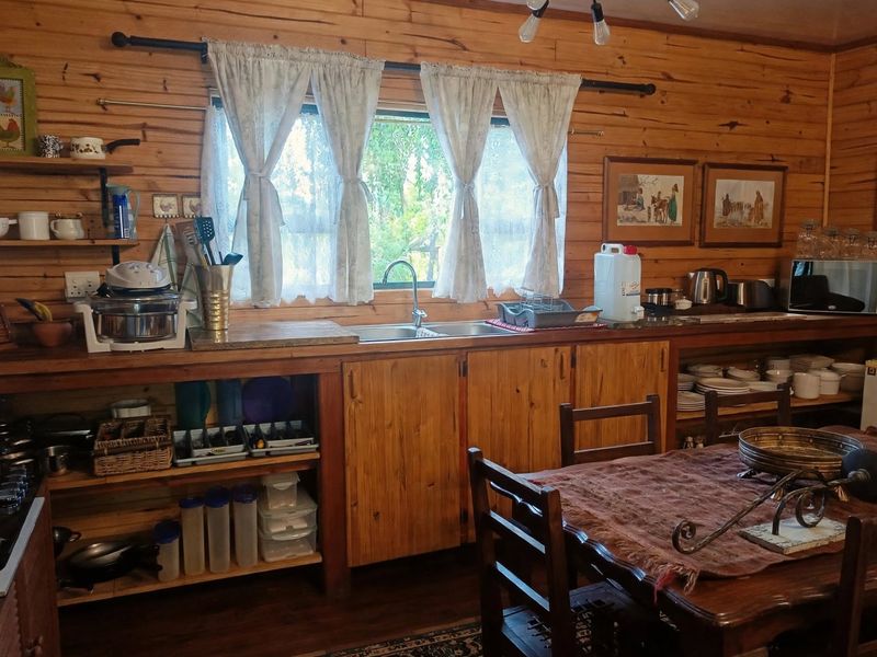 Rustic cottage kitchen featuring timber wall paneling, custom wooden cabinetry, open shelving, and a stainless steel sink with a view of the garden.