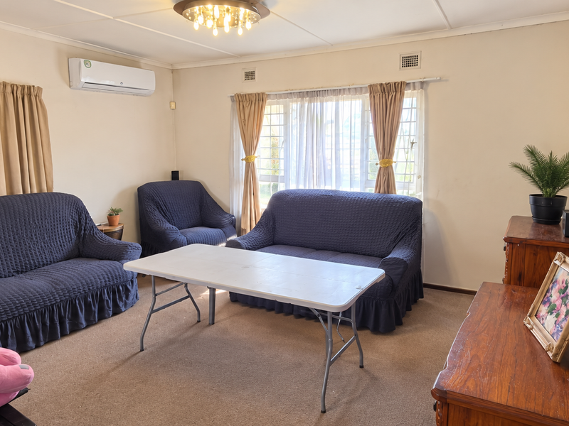 A comfortable living room featuring a wall-mounted air conditioning unit, carpeted flooring, and large windows with security bars.