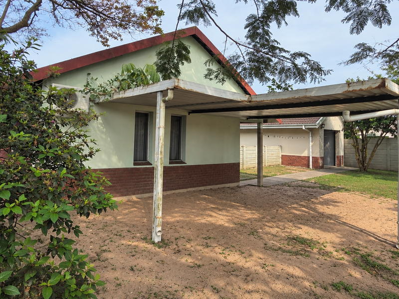 Rear exterior view of the property featuring a metal carport, brick-base facade, and a concrete boundary wall in a garden setting.