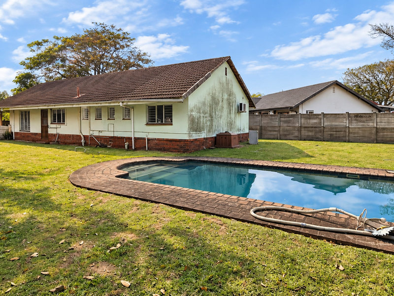 Rear view of the property featuring a swimming pool with brick coping, a large lawn, and a tiled roof structure with visible exterior wall staining.