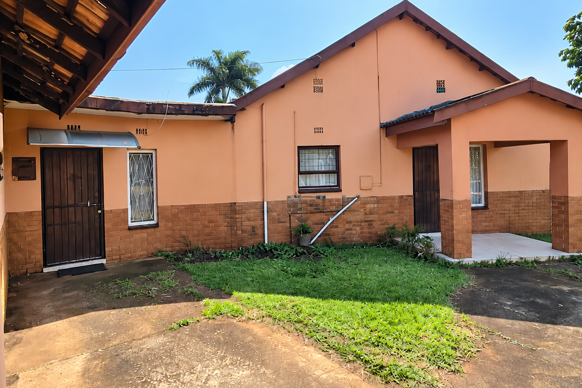 Rear view of the house featuring a brick and plaster facade, covered patio area, and concrete driveway with established garden surroundings.