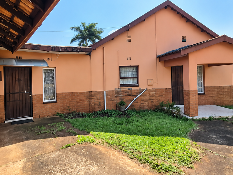 Rear view of the house featuring a brick and plaster facade, covered patio area, and concrete driveway with established garden surroundings.