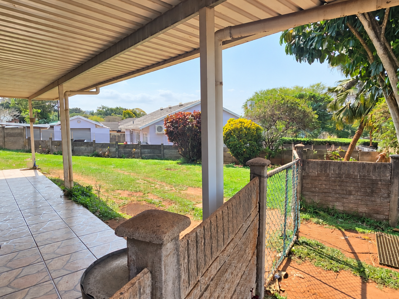 A covered outdoor patio area featuring tiled flooring and support pillars, overlooking a spacious backyard with established trees and perimeter fencing.