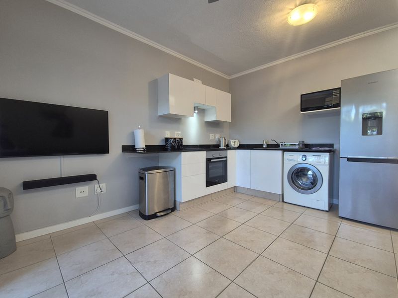 Compact open-plan kitchen featuring modern white cabinetry, dark stone countertops, built-in oven, and integrated laundry facilities with tiled flooring.