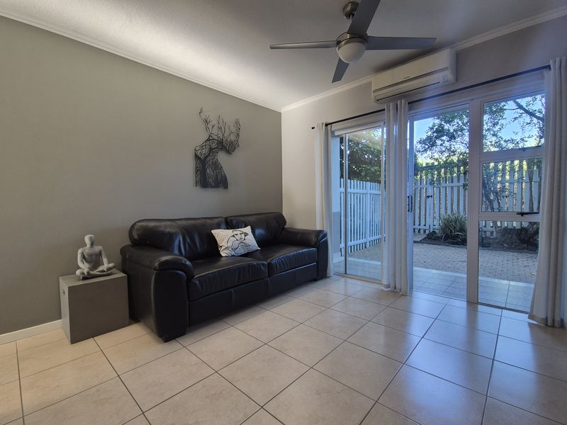 A bright living room featuring tiled flooring, a ceiling fan, a wall-mounted air conditioner, and sliding glass doors leading to an outdoor patio area.