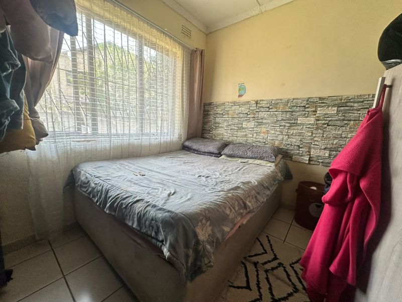 A compact bedroom featuring tiled flooring, a stone-effect feature wall, and a large window with security bars.