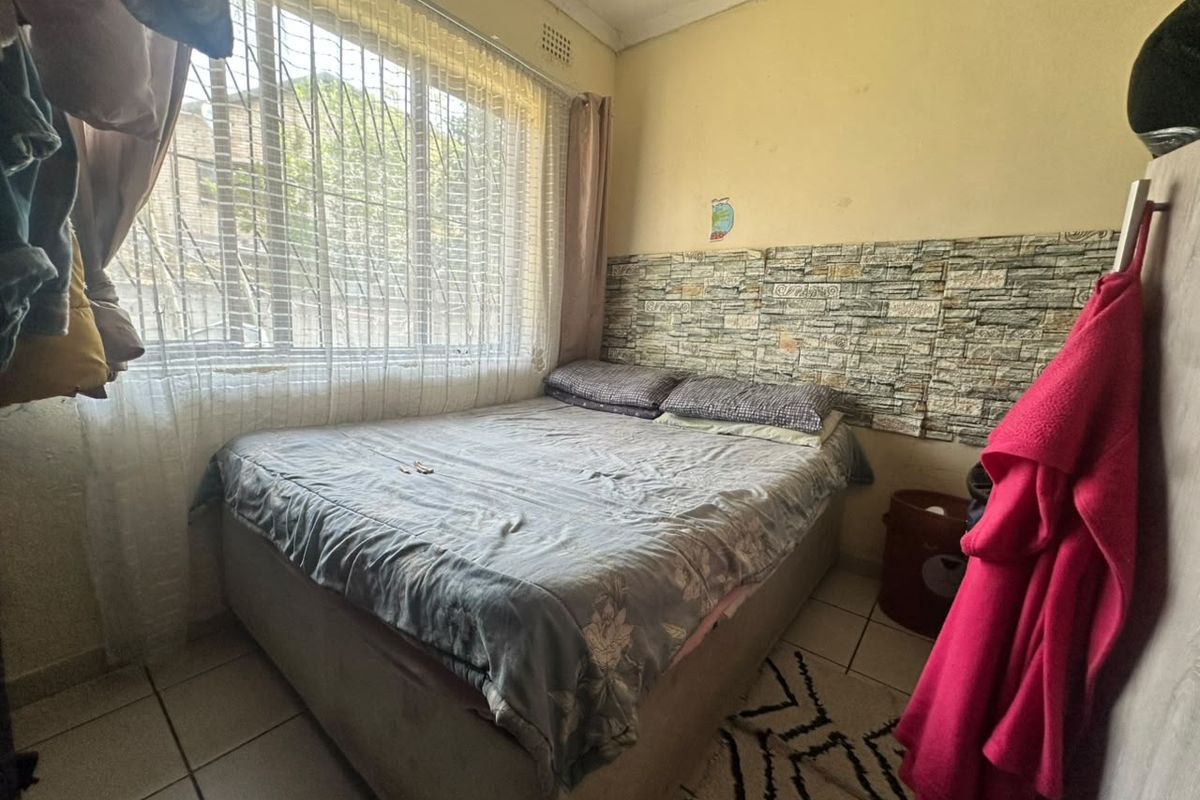 A compact bedroom featuring tiled flooring, a stone-effect feature wall, and a large window with security bars.