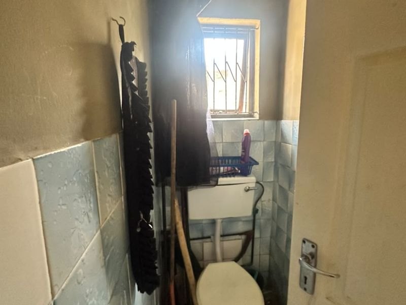 A compact, dated bathroom featuring a standard toilet, ceramic wall tiling, and a small window with security bars.