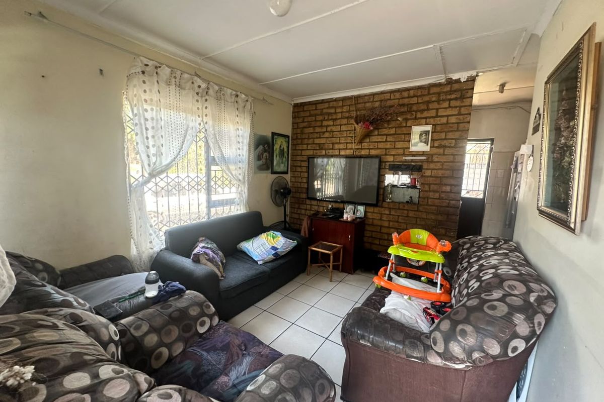 A living room featuring an exposed brick accent wall, ceramic tiled flooring, and security gate access, requiring cosmetic updates to improve its overall condition.