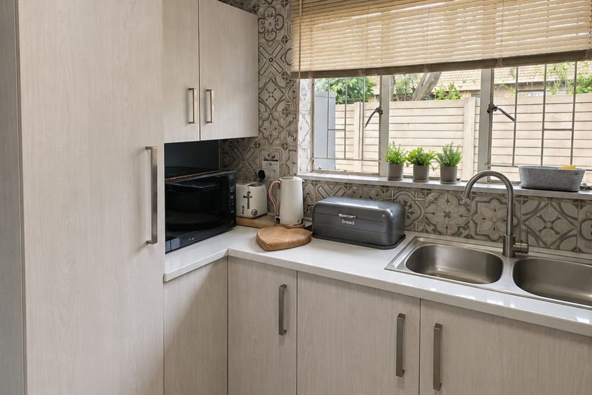 A functional kitchen featuring light wood-grain cabinetry, a double stainless steel sink, patterned tile backsplash, and neutral floor tiles.