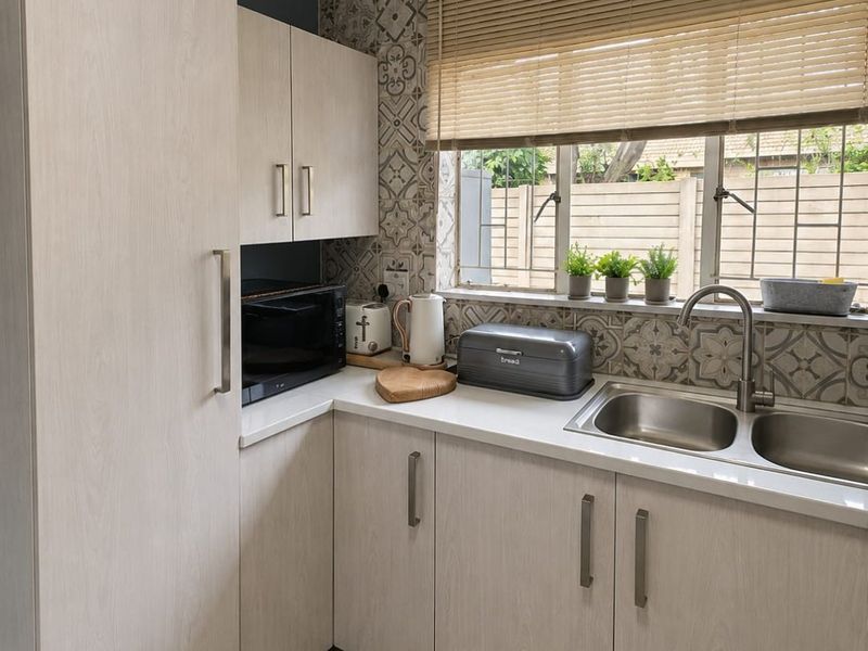 A functional kitchen featuring light wood-grain cabinetry, a double stainless steel sink, patterned tile backsplash, and neutral floor tiles.