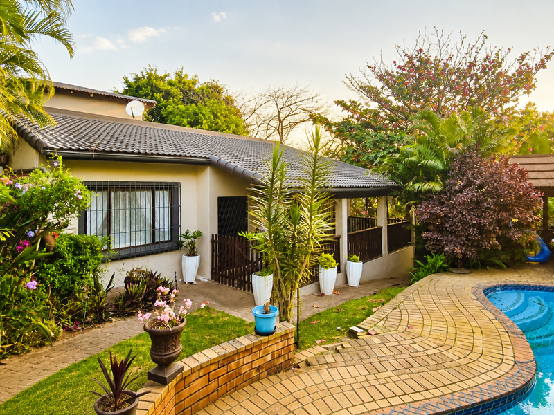 A well-maintained outdoor pool area featuring brick paving, a covered patio, and established garden landscaping surrounding the swimming pool.